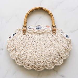 May include: A cream-colored crochet handbag shaped like a seashell with a scalloped edge. The bag features a natural bamboo handle attached with tan leather tabs and silver rivets. The interior lining is white fabric with a blue seashell print.