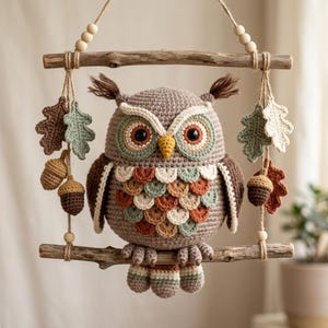Op de afbeelding: Een gehaakte uil wandhanger opgehangen aan twee horizontale houten takken. De uil heeft bruine, crème en saliegroene veren, met bungelende gehaakte eikenbladeren en eikels aan jute touw, geaccentueerd door kleine houten kralen.