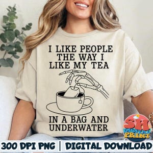 May include: Beige t-shirt with the text "I LIKE PEOPLE THE WAY I LIKE MY TEA IN A BAG AND UNDERWATER." The graphic shows a skeleton hand placing a tea bag into a teacup. The words "DIGITAL PRINT" are also visible.