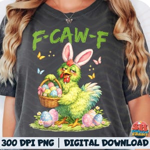 Puede incluir: Camiseta gris oscuro con un dibujo de un pollo de dibujos animados con orejas de conejo y una cesta de huevos de Pascua. La camiseta tiene el texto "F-CAW-F" en verde. También aparecen mariposas y huevos de Pascua.