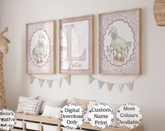 May include: Three framed prints with a duck illustration in a nursery setting. The prints feature a duck in a floral frame, a letter "L" with the name "Lottie", and a duck in a floral frame. The prints are in a light pink and beige color scheme.