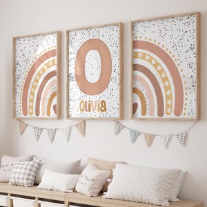 May include: Three framed wall art prints with a rainbow design in shades of peach, tan, and gold. The centre print features the letter "O" and the name "Olivia". The prints are displayed above a bench with pillows and a decorative banner.