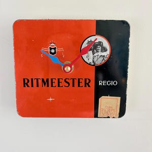 May include: A rectangular wall clock featuring a split orange and black design. The text RITMEESTER REGIO is printed on the front. It includes a circular graphic of a person in a hat, a blue hour hand, a red minute hand, and a small beige tax stamp.