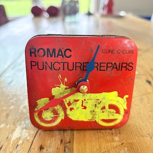 May include: A square red metal clock featuring the black text ROMAC PUNCTURE REPAIRS and CURE-C-CURE. A yellow graphic of a motorcycle is printed on the front. The clock has a blue hour hand, a red minute hand, and a gold-colored center cap.