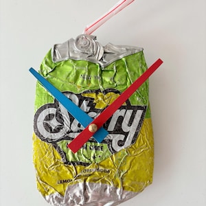 May include: A unique clock made from a crushed, repurposed soda can. The clock face features the "Cherry" logo, with blue and red hands indicating the time. A red and white striped straw is attached to the top.