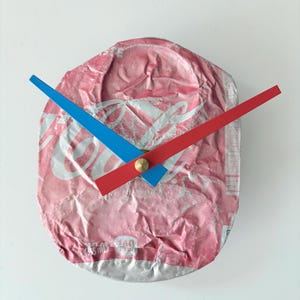 May include: A unique wall clock made from a crushed pink soda can. The clock features red and blue hands, a brass center, and a white background. The clock's hands are positioned to show the time.