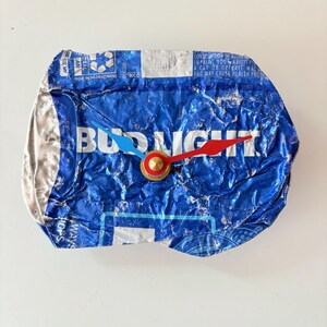 May include: A unique wall clock crafted from a crushed Bud Light beer can. The clock face is blue with the Bud Light logo visible. It features red and blue clock hands and a gold-colored center.