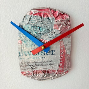 Op de afbeelding: Een unieke wandklok gemaakt van een hergebruikte Budweiser bierblikje. De klok heeft de originele graphics van het blikje in zilver, rood en blauw. De wijzers zijn rood en blauw, met een gouden middelpunt. De klok leest "Budweiser" en "Lager Beer".