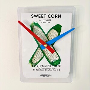May include: A rectangular wall clock featuring a vintage seed packet design with the words "SWEET CORN" and an illustration of corn. The clock has red and blue hands and is encased in a clear plastic frame.