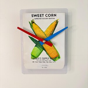 May include: A wall clock featuring a vintage "Sweet Corn" graphic with crossed corn cobs and green leaves. The clock has red and blue hands and includes the text "Farmer's Supply House".
