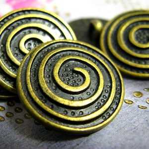 Pode incluir: Três botões de metal dourados com um design de espiral preto. Os botões têm uma superfície texturizada e um padrão de espiral em relevo.