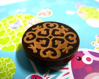 May include: A round wooden button with a brown and black geometric design. The design features a repeating pattern of hearts and swirls.