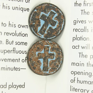 Puede incluir: Dos botones redondos de cerámica con un diseño de cruz azul sobre fondo marrón. Los botones tienen un aspecto desgastado y envejecido.