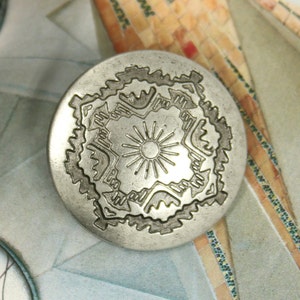 Könnte beinhalten: Eine silberfarbene Metallnadel mit einem geometrischen Design. Das Design zeigt eine zentrale Sonnenstrahlen umgeben von einem sich wiederholenden Muster aus stilisierten Formen.