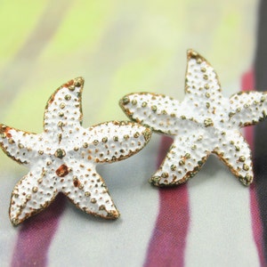 Puede incluir: Dos pendientes de estrella de mar blancos con detalles dorados. Las estrellas de mar tienen una superficie texturizada y son una tendencia popular de joyería de verano.