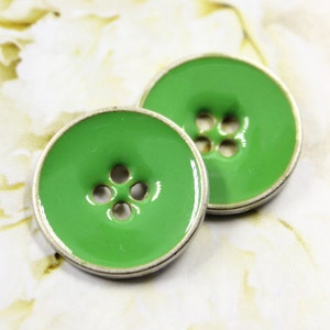 Può includere: Due bottoni rotondi verde brillante con bordo color argento. Ogni bottone ha quattro fori al centro per cucire. I bottoni sono posti su uno sfondo floreale morbido.
