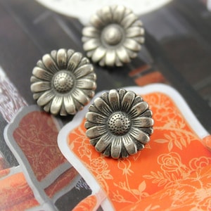 May include: Three silver metal daisy-shaped buttons with a raised center. The buttons are on a patterned orange and white surface.