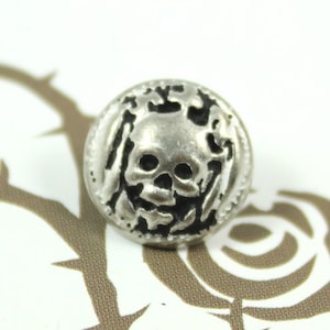Könnte beinhalten: Silberfarbener Knopf mit einem schwarzen Totenkopf-Design. Der Totenkopf ist detailliert mit Kreuzknochen und anderen dekorativen Elementen gestaltet.