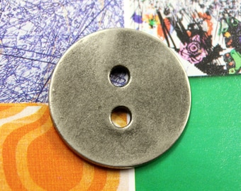 May include: A circular, silver button with two holes. The button has a textured surface and is set against a colourful background with orange, green, and purple patterns. Suitable for sewing projects.
