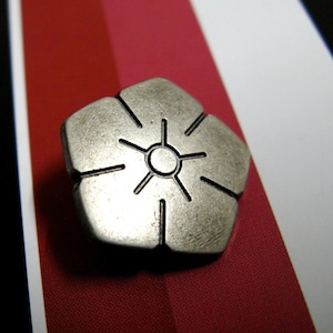 Puede incluir: Un pin de metal plateado en forma de flor con un diseño de sol estilizado en el centro. El pin está sobre un fondo de rayas rojas y blancas.