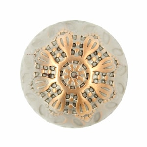 Puede incluir: Un botón redondo de color plateado con un diseño en relieve de color cobre de una flor estilizada. La flor tiene un centro circular con un círculo concéntrico más pequeño en el centro.