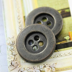 Peut inclure: Deux gros boutons métalliques ronds de couleur gris foncé avec quatre trous chacun. Les boutons ont un aspect légèrement usé, antique.