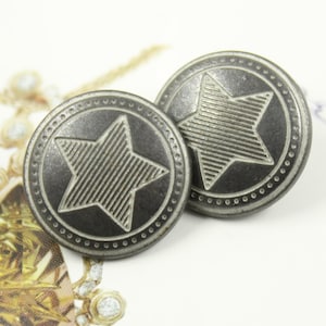 May include: Two silver-toned metal buttons with a star design. The star has a textured, striped pattern. The buttons have a circular, beaded border.