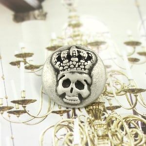May include: Silver-toned button with a skull design wearing a crown. The button is round and has a textured surface.