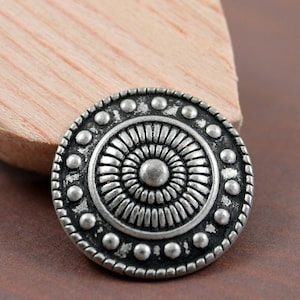 Puede incluir: Botón de metal plateado con un diseño circular que presenta un patrón de círculo concéntrico elevado y un borde exterior con cuentas.