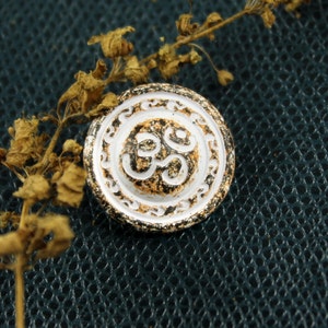 Pode incluir: Um botão circular branco com uma borda dourada e um símbolo Om preto no centro. O botão está sobre uma superfície texturizada azul escura.