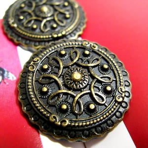 Puede incluir: Dos botones de metal de color dorado antiguo con diseños intrincados. Los botones tienen una forma circular con un centro elevado y un borde decorativo.
