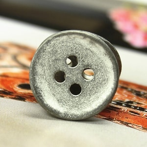 Pode incluir: Close-up de um botão metálico desgastado com quatro orifícios. O botão tem uma aparência vintage e envelhecida, com uma textura ligeiramente áspera e uma cor prateada suave. O botão é provavelmente para roupas ou projetos de artesanato.