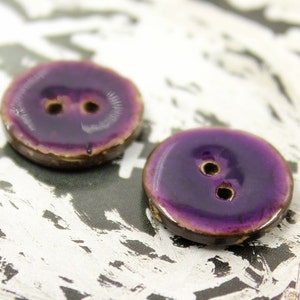 Peut inclure: Deux boutons en céramique violet avec deux trous chacun. Les boutons ont un bord légèrement surélevé et une finition brillante.
