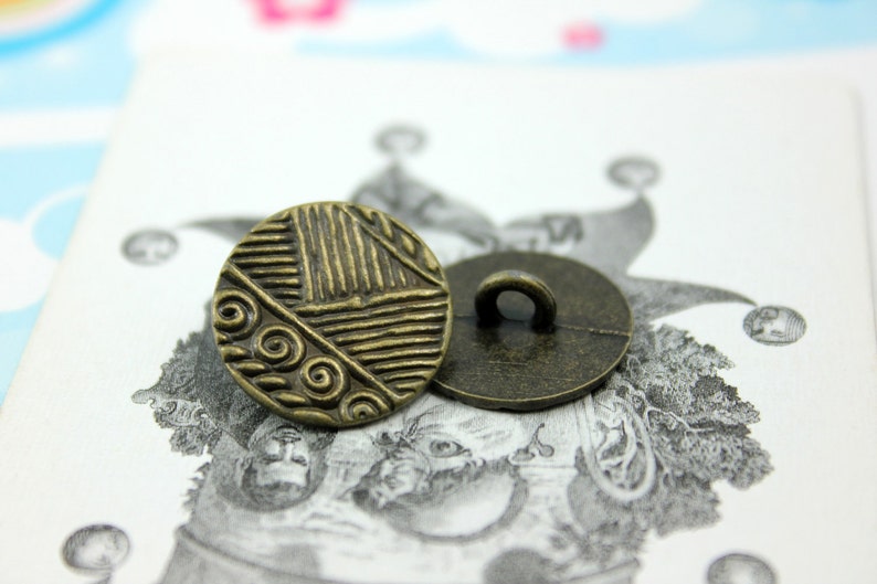 Puede incluir: Dos botones de color dorado antiguo con un dise&ntilde;o decorativo. Un bot&oacute;n tiene un dise&ntilde;o circular con un patr&oacute;n de espiral y el otro bot&oacute;n es liso.