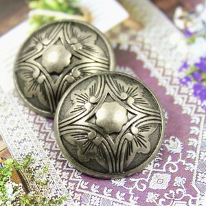 May include: Two silver-toned metal buttons with a decorative floral design. The buttons have a raised centre and a slightly textured surface.