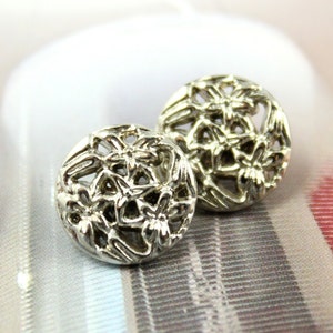 Puede incluir: Dos pendientes de metal plateados con un diseño floral. Los pendientes son redondos y tienen un patrón delicado e intrincado.