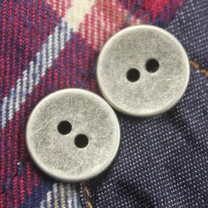 May include: Two silver buttons with two holes each, set against a red and blue plaid fabric.