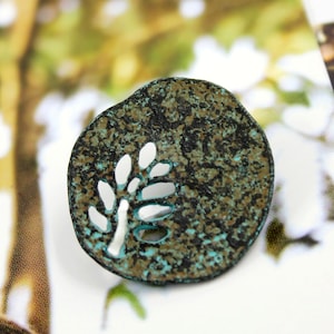 May include: A round, rustic, turquoise and brown metal charm with a cut-out design of a branch with leaves.