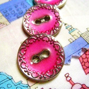 May include: Three pink buttons with silver metal rims and ornate designs. The buttons have a shiny, glossy finish and a single hole in the center.