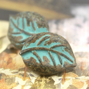 Puede incluir: Dos botones en forma de hoja de color azul turquesa con un acabado marrón rústico. Los botones tienen un diseño de hoja en relieve y un aspecto desgastado.