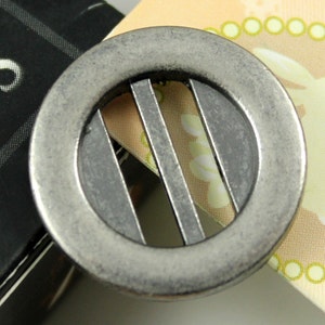 May include: A silver metal button with a circular design and three vertical bars in the center.