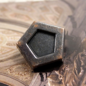 Puede incluir: Un botón metálico desgastado en forma de pentágono, con una forma interior empotrada de cinco lados. El botón tiene un aspecto oscuro y envejecido con toques de bronce, lo que sugiere una estética antigua o vintage. El botón es probablemente para ropa o manualidades.