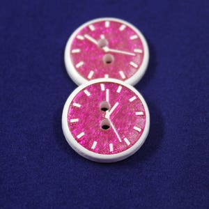 Puede incluir: Dos botones de color rosa y blanco en forma de reloj con manecillas blancas y un borde blanco. Los botones tienen un acabado brillante.