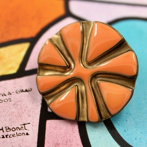 May include: A peach-colored enamel flower brooch with a gold-toned metal frame. The flower has six petals and a center that is slightly raised. The brooch is a vintage piece and has a unique design.