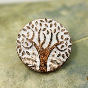 May include: A round, white ceramic button with a brown tree design. The tree has a thick trunk and branches with leaves.