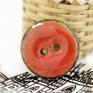Può includere: Un bottone in ceramica rosso con un bordo marrone e due fori. Il bottone ha una finitura lucida.