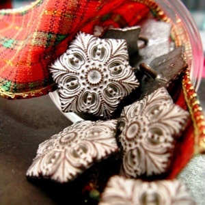 May include: A collection of decorative metal snowflake ornaments in various sizes. The ornaments are white with intricate designs and are arranged on a red and green plaid fabric.