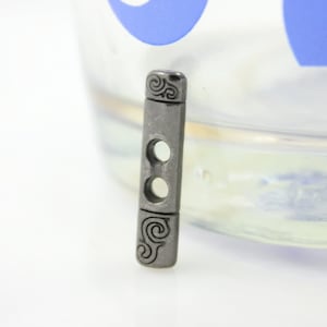 Könnte beinhalten: Ein silberner Metallknopf mit einem dekorativen Wirbelmuster. Der Knopf hat zwei Löcher zum Nähen.