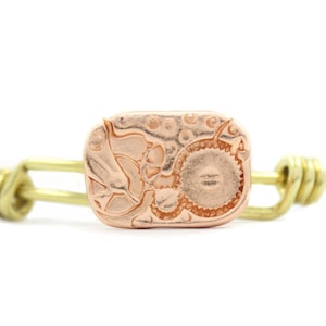 Puede incluir: Un brazalete de brazalete de latón con un dije de oro rosa adornado con un diseño detallado e intrincado de engranajes y otros elementos mecánicos.