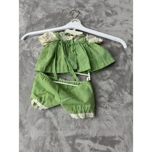 Può includere: Un completo per bambola a quadretti vichy verdi e bianchi composto da una camicetta a maniche corte con motivo floreale e pantaloncini a sbuffo abbinati. La camicetta presenta un bordo in pizzo bianco sulle maniche e i pantaloncini sul fondo.